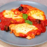 Delizioso pollo alla pizzaiola su un piatto grigio, con pomodoro, mozzarella filante, olive nere e una foglia di basilico.