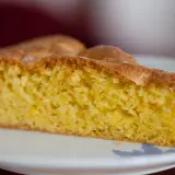 Una fetta di torta alle mandorle dorata e soffice, con una crosta leggermente caramellata, servita su un piatto bianco.