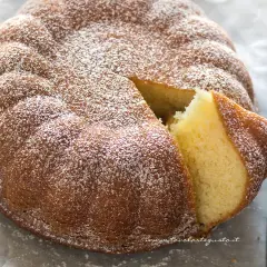 Una soffice torta allo yogurt, cosparsa di zucchero a velo, con una fetta tagliata che ne rivela l'interno dorato e invitante.