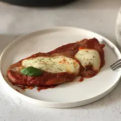 Scaloppine alla pizzaiola su un piatto bianco, con mozzarella filante, salsa di pomodoro e una foglia di basilico.
