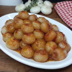 Cipolline in agrodolce, caramellate e lucide, servite su un piatto bianco. Sullo sfondo, cipolle fresche e un tovagliolo a quadri rossi.