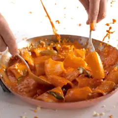 Mani che mescolano paccheri alla Vittorio in padella, con salsa arancione che schizza.