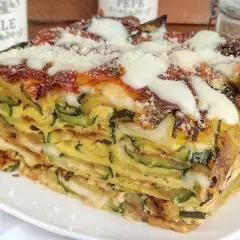 Una fetta di timballo di zucchine stratificato, con formaggio fuso e parmigiano grattugiato, servito su un piatto bianco.