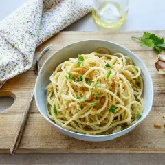 Un piatto di spaghetti con la mollica croccante, guarnito con prezzemolo fresco. Accanto, aglio e un bicchiere d'acqua.