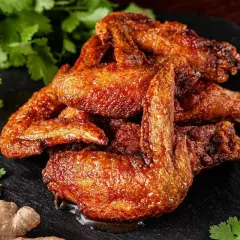 Un mucchio di ali di pollo dorate e succulente, condite con spezie, su una pietra scura, con coriandolo, zenzero e aglio sullo sfondo.