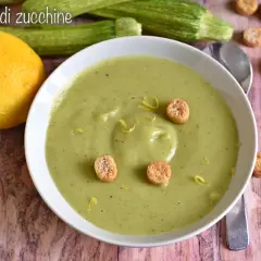 Vellutata di zucchine cremosa, guarnita con crostini e scorza di limone. Accanto, zucchine fresche e un limone intero.