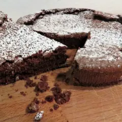 Una fetta di tenerina al cioccolato, cosparsa di zucchero a velo, è stata appena tagliata da una torta intera su un tagliere di legno.