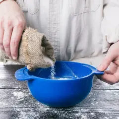 Mani versano zucchero da un sacchetto di juta in una ciotola blu, preparando ingredienti per pasta di zucchero.