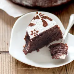 Una fetta di torta caprese bimby, soffice e al cioccolato, decorata con zucchero a velo e cacao, su un piatto bianco a forma di cuore.