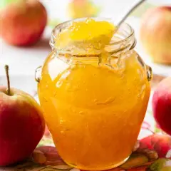 Un barattolo di marmellata di mele dorata, con una cucchiaiata che ne assaggia la dolcezza, circondato da mele rosse fresche.