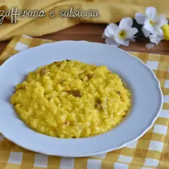 Un cremoso risotto salsiccia e zafferano, servito su un piatto bianco, con fiori gialli e bianchi sullo sfondo.