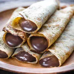 Un mucchio di crepes arrotolate, ripiene di golosa Nutella, su un piatto di legno.