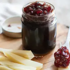 Un vasetto di marmellata di cipolle rosse, con un cucchiaino che ne raccoglie una porzione, accanto a fette di formaggio su un tagliere di legno.