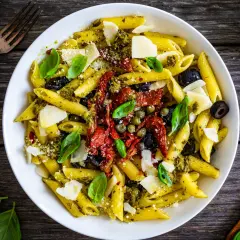 Penne con pesto, pomodori secchi, olive nere, capperi e scaglie di parmigiano. Un piatto ricco di sapori mediterranei.