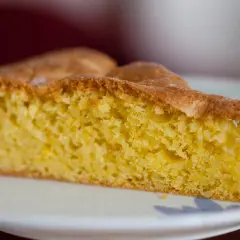 Una fetta di torta alle mandorle dorata e soffice, con una crosta leggermente caramellata, servita su un piatto bianco.