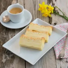 Tre fette di torta magica ricetta, cremosa e dorata, su un piatto bianco, con caffè e narcisi.