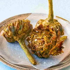 Due carciofi alla giudia dorati e croccanti, cosparsi di sale grosso, su un vassoio d'argento.
