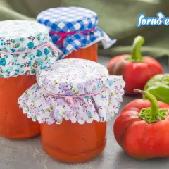 Barattoli di crema di peperoni fatti in casa, decorati con stoffe floreali e a quadretti, accanto a peperoni freschi.