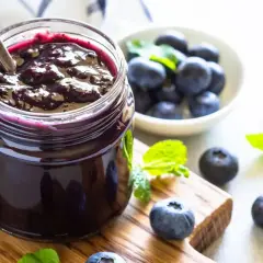 Vasetto di marmellata di mirtilli fatto in casa, con mirtilli freschi e foglie di menta su un tagliere di legno.