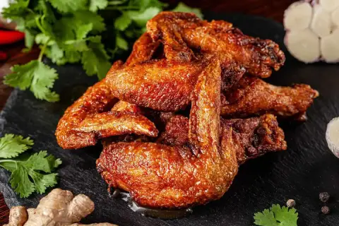 Un mucchio di ali di pollo dorate e succulente, condite con spezie, su una pietra scura, con coriandolo, zenzero e aglio sullo sfondo.