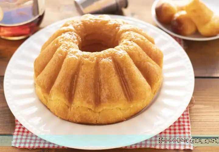 Un soffice babà a ciambella, lucido di bagna, pronto per essere gustato. Accanto, altri babà e una bottiglia di rum.