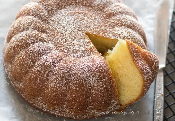 Una soffice torta allo yogurt, cosparsa di zucchero a velo, con una fetta tagliata che ne rivela l'interno dorato e invitante.