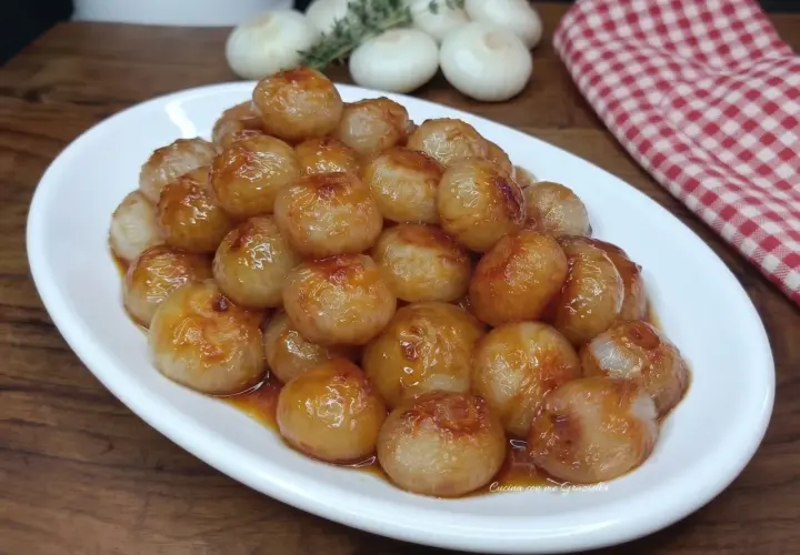 Cipolline in agrodolce, caramellate e lucide, servite su un piatto bianco. Sullo sfondo, cipolle fresche e un tovagliolo a quadri rossi.