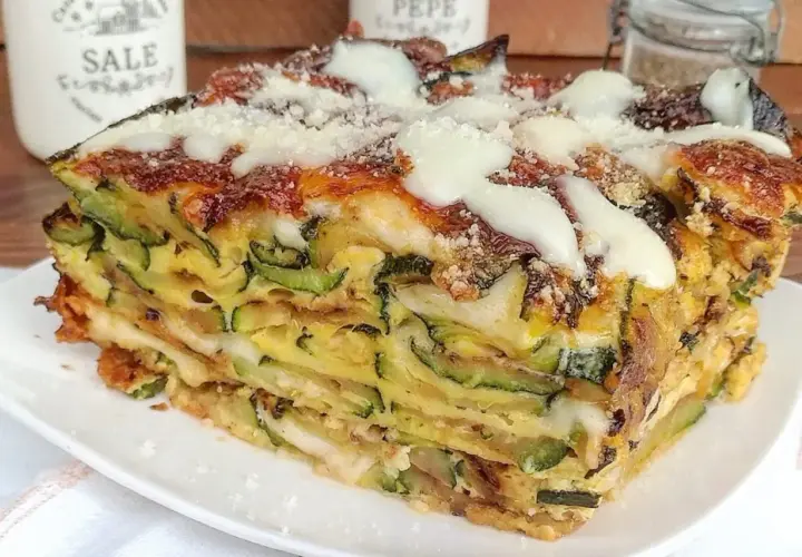 Una fetta di timballo di zucchine stratificato, con formaggio fuso e parmigiano grattugiato, servito su un piatto bianco.