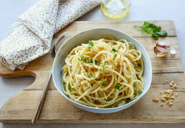 Un piatto di spaghetti con la mollica croccante, guarnito con prezzemolo fresco. Accanto, aglio e un bicchiere d'acqua.
