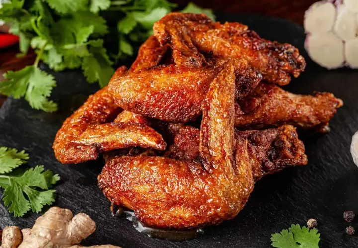 Un mucchio di ali di pollo dorate e succulente, condite con spezie, su una pietra scura, con coriandolo, zenzero e aglio sullo sfondo.