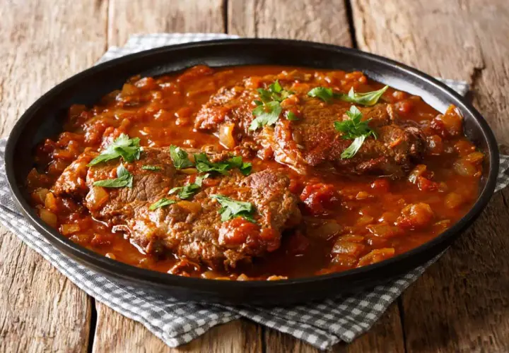 Carne alla pizzaiola morbida in un piatto scuro, con pomodoro e prezzemolo fresco.