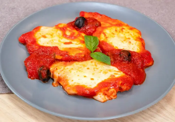 Delizioso pollo alla pizzaiola su un piatto grigio, con pomodoro, mozzarella filante, olive nere e una foglia di basilico.