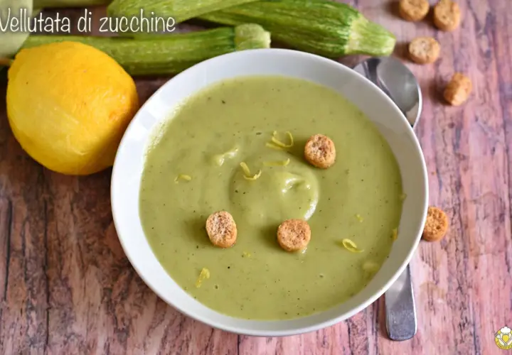Vellutata di zucchine cremosa, guarnita con crostini e scorza di limone. Accanto, zucchine fresche e un limone intero.