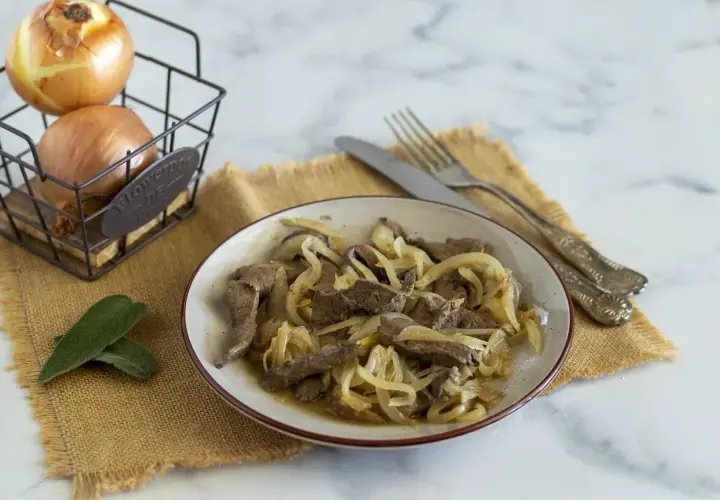 Piatto di fegato alla veneziana con cipolle dorate, accompagnato da due foglie di salvia e cipolle in un cestino.
