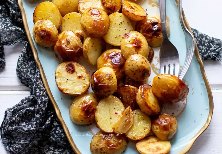 Patate novelle al forno dorate e croccanti, servite su un piatto azzurro con forchette.