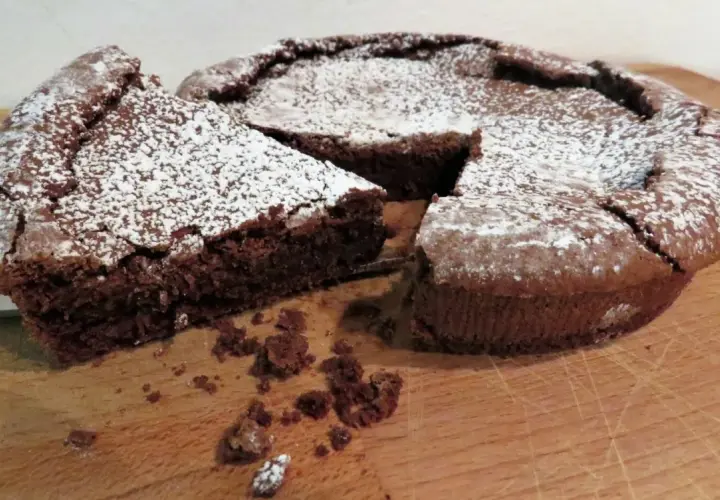 Una fetta di tenerina al cioccolato, cosparsa di zucchero a velo, è stata appena tagliata da una torta intera su un tagliere di legno.