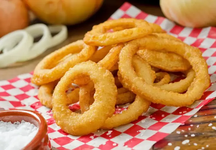 Anelli di cipolla dorati e croccanti, preparati con una **pastella per fritti** perfetta, serviti su carta a quadri rossi con sale grosso.