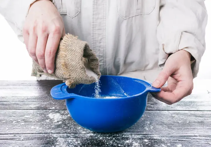 Mani versano zucchero da un sacchetto di juta in una ciotola blu, preparando ingredienti per pasta di zucchero.