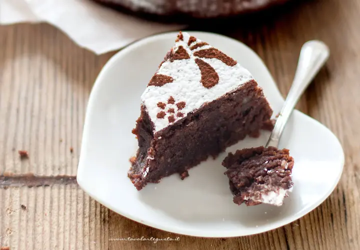 Una fetta di torta caprese bimby, soffice e al cioccolato, decorata con zucchero a velo e cacao, su un piatto bianco a forma di cuore.