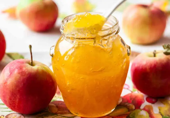 Un barattolo di marmellata di mele dorata, con una cucchiaiata che ne assaggia la dolcezza, circondato da mele rosse fresche.