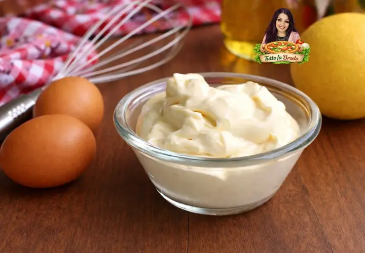 Una ciotola di cremosa maionese fatta in casa, con uova, limone e frusta sullo sfondo.