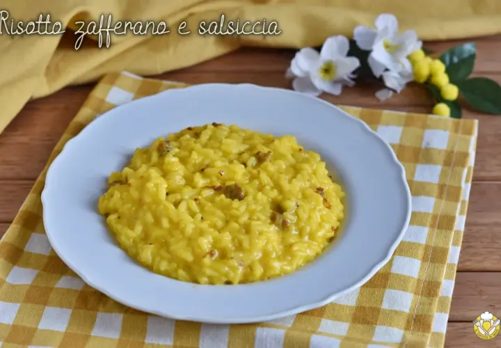 Un cremoso risotto salsiccia e zafferano, servito su un piatto bianco, con fiori gialli e bianchi sullo sfondo.