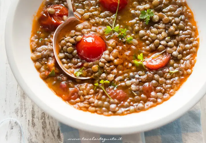 Una ciotola di zuppa di lenticchie fumante, con pomodorini e prezzemolo fresco. Un cucchiaio raccoglie una porzione generosa di questo piatto confortante.