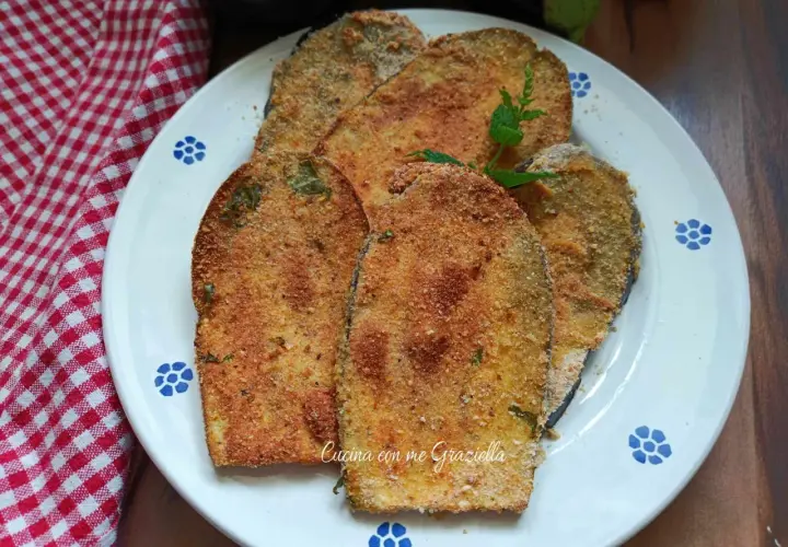 Fette dorate di cotoletta di melanzane impanate, guarnite con menta fresca, su un piatto bianco con decori blu.