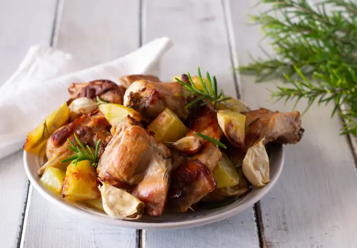 Piatto invitante di coniglio al forno con patate arrosto, aglio e rosmarino su un tavolo di legno.