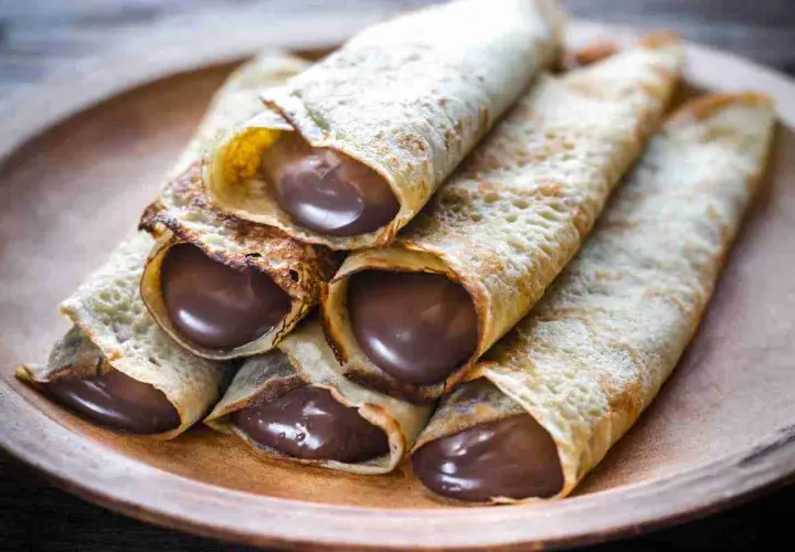 Un mucchio di crepes arrotolate, ripiene di golosa Nutella, su un piatto di legno.