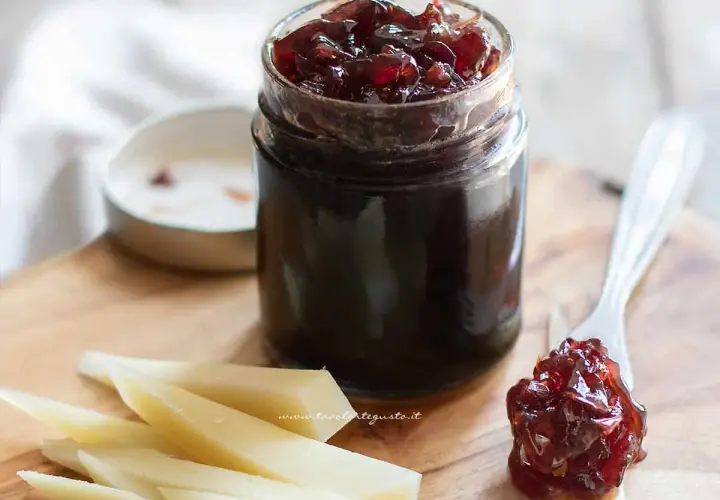 Un vasetto di marmellata di cipolle rosse, con un cucchiaino che ne raccoglie una porzione, accanto a fette di formaggio su un tagliere di legno.