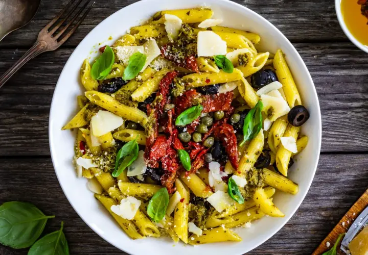 Penne con pesto, pomodori secchi, olive nere, capperi e scaglie di parmigiano. Un piatto ricco di sapori mediterranei.