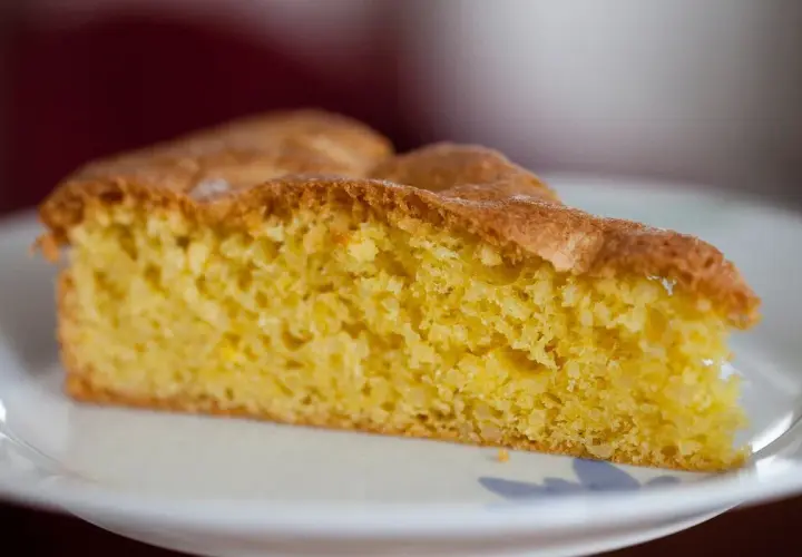 Una fetta di torta alle mandorle dorata e soffice, con una crosta leggermente caramellata, servita su un piatto bianco.