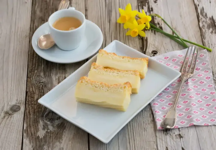 Tre fette di torta magica ricetta, cremosa e dorata, su un piatto bianco, con caffè e narcisi.